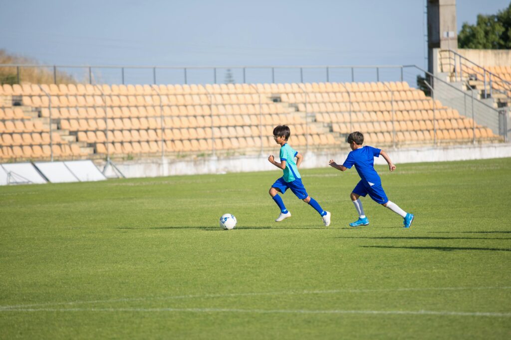 Two boys energetically play soccer on a sunny day in an empty stadium field.